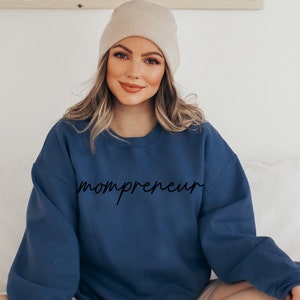 May include: A navy blue crew neck sweatshirt with the word "mompreneur" in a cursive font. The model is wearing a beige beanie. The sweatshirt is a casual style.