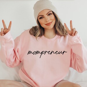 May include: A pale pink sweatshirt with the word "mompreneur" in black script. The model is wearing a beige beanie and making a peace sign with their fingers.