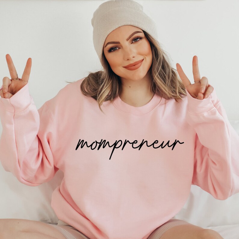 May include: A pale pink sweatshirt with the word "mompreneur" in black script. The model is wearing a beige beanie and making a peace sign with their fingers.