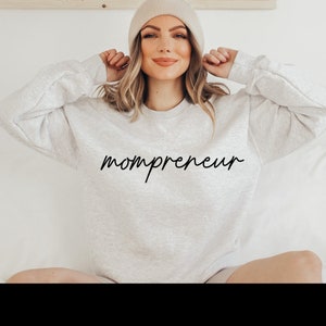 May include: A light grey sweatshirt with the word "mompreneur" in black script. The model is wearing a beige beanie. The sweatshirt is a casual style, suitable for everyday wear.