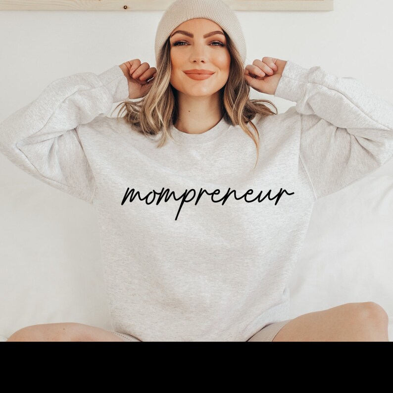 May include: A light grey sweatshirt with the word "mompreneur" in black script. The model is wearing a beige beanie. The sweatshirt is a casual style, suitable for everyday wear.