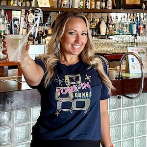 May include: A woman in a navy blue t-shirt with the text "TUNE-IN LOUNGE" and a black skirt holds a cocktail. The bar has vintage televisions and a sign that says "Order Here".
