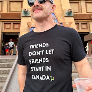 May include: A black short-sleeved t-shirt featuring the white text "FRIENDS DON'T LET FRIENDS START IN CANADA" with a small colourful cocktail graphic next to the word Canada, worn with a brown baseball cap and black sunglasses.