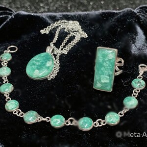 May include: A jewelry set featuring a silver necklace with a teardrop pendant, a bracelet, and a ring. The jewelry has green gemstones. The ring has a rectangular gemstone. The bracelet and necklace have round gemstones.