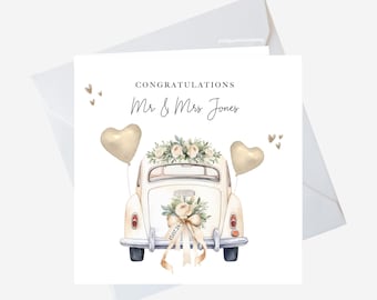 May include: White wedding card with a white vintage car with gold heart balloons, floral decorations, and a ribbon with the date "18.07.24" written on it. The text "Congratulations Mr & Mrs Jones" is written above the car.