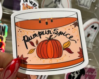 May include: A die-cut sticker featuring a cylindrical candle with orange wax and three lit wicks. The label displays the text Pumpkin Spice in black cursive script above a central orange pumpkin illustration surrounded by small colorful autumn leaves.