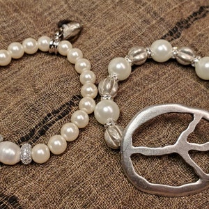Può includere: Due braccialetti di perle bianche, uno con un ciondolo a forma di segno della pace in argento. I braccialetti sono su uno sfondo di tessuto marrone.