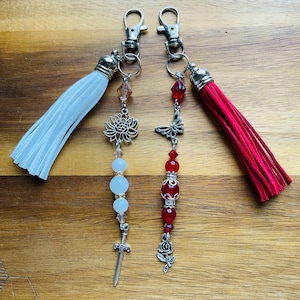 May include: Two keychains with white and red tassels. The white keychain has a silver charm of a flower, a sword, and white beads. The red keychain has a silver charm of a butterfly, a rose, and red beads.