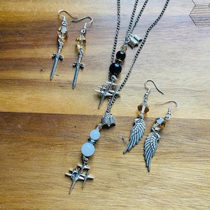 Puede incluir: Una colección de joyas plateadas sobre una superficie de madera. El conjunto incluye dos pares de pendientes, uno con colgantes de espada y otro con colgantes de ala. Un collar con múltiples cadenas presenta detalles de estrella, libro y cuentas.