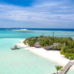 Puede incluir: Vista aérea de una isla tropical con playas de arena blanca, agua turquesa y exuberante vegetación verde. Hay varios edificios de madera en la isla, incluida una cabaña con techo de paja y una plataforma de madera.