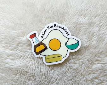 May include: A white sticker with a cartoon illustration of a fried egg, a small bowl of rice, a stick of butter, and a bottle of soy sauce. The text "Asian Kid Breakfast" is written in a circle around the food.