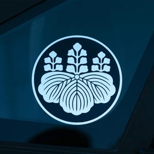 Puede incluir: Una calcomanía circular blanca con un diseño de escudo floral estilizado. El escudo se compone de tres elementos florales más pequeños sobre tres hojas más grandes en forma de abanico, todo encerrado dentro de un círculo blanco. Fondo azul oscuro.