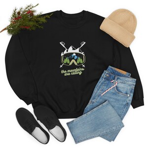 Könnte beinhalten: Schwarzer Pullover mit einer weißen Grafik von Skibrillen mit einer Bergszene im Inneren. Der Text "the mountains are calling" befindet sich unter den Brillen.