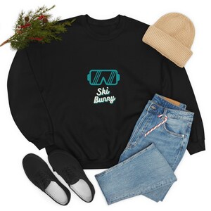 Könnte beinhalten: Schwarzes Sweatshirt mit einer türkisfarbenen Skibrillen-Grafik und dem Text "Ski Bunny". Das Bild zeigt auch eine beige Mütze, blaue Jeans und schwarze Schuhe. Ein Zweig mit Grünzeug befindet sich in der oberen linken Ecke.