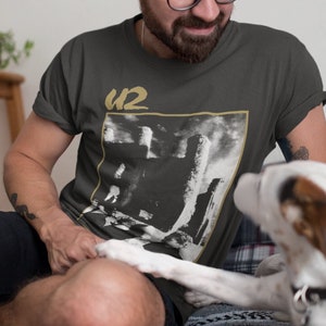 以下が含まれることがあります： 黒と白のU2のバンドの画像とバンド名が金色の文字で書かれた濃いグレーのTシャツを着た人。白とグレーの模様の毛布のあるベッドに座っています。白と茶色の犬がその人の隣に座ってカメラを見ています。