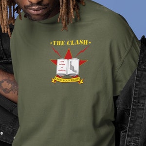 May include: Olive green t-shirt featuring a red star with the text "THE CLASH" above and "KNOW YOUR RIGHTS" below. The graphic includes a book with the words "THE FUTURE IS UNWRITTEN".