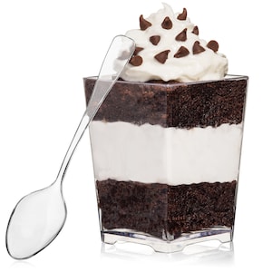 May include: A clear plastic dessert cup filled with layers of chocolate cake and white frosting, topped with whipped cream and chocolate chips. A clear plastic spoon is next to the cup.