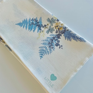 May include: White linen tea towel with a blue and green floral design. The towel has the text "LITTLE" and a heart logo in the bottom right corner.