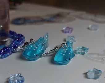 Pode incluir: Um par de brincos de tubarão-baleia azul com manchas brancas. Os brincos são feitos de plástico transparente e têm ganchos prateados.