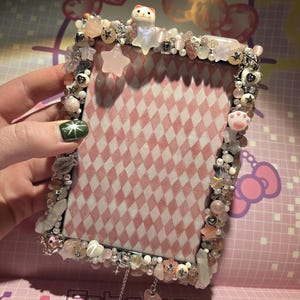 Pode incluir: Uma moldura decorativa com um fundo de padrão de losangos rosa e branco. A moldura é adornada com contas, cristais e berloques em tons de rosa, branco e prata. Uma pequena figura de gato e berloques em forma de estrela adornam o topo.