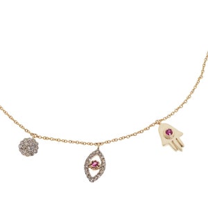 May include: A gold chain necklace with three charms: a hamsa hand, an evil eye, and a round charm with small diamonds. Each charm has a small pink gemstone in the center.