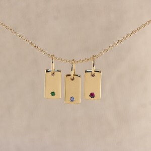 Könnte beinhalten: Eine goldene Halskette mit drei rechteckigen Anhängern, die jeweils mit einem kleinen Edelstein besetzt sind. Die Edelsteine sind grün, blau und rot. Die Anhänger hängen an einer feinen Goldkette.