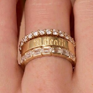 May include: A stack of three gold rings. The top ring features a row of small round clear stones. The middle band is smooth gold with the text 'til death' engraved in a gothic font. The bottom ring features a row of rectangular clear stones.