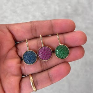 May include: Three circular pendants featuring a gold-toned metal frame and a surface densely covered in small, sparkling stones. One pendant is blue, one is magenta, and one is green, each attached to a thin, vertical gold-toned metal bail.