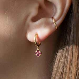 May include: A gold hoop earring with a small, four-leaf clover charm adorned with red gemstones. A second gold earring is visible on the ear. The jewelry is set against the backdrop of a person's ear and hair.