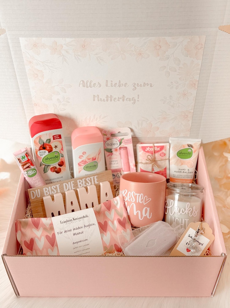 Może przedstawiać: R&oacute;żowe pudełko prezentowe wypełnione artykułami do pielęgnacji. Zawiera kubek z napisem "Najlepsza mama", świecę, balsamy, balsam do ust i drewniany znak z napisem "Jesteś najlepszą mamą". Pudełko jest ozdobione kwiatowymi wzorami.