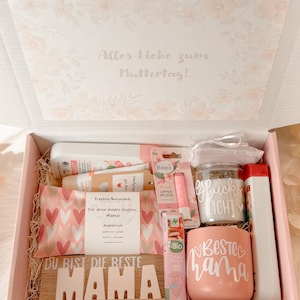 Może przedstawiać: R&oacute;żowe pudełko prezentowe wypełnione produktami do pielęgnacji. Zawiera kubek z napisem "BESTE MAMA", drewniany znak z napisem "DU BIST DIE BESTE MAMA" i inne produkty kosmetyczne. Pudełko jest ozdobione kwiatowymi wzorami i niemieckim tekstem.