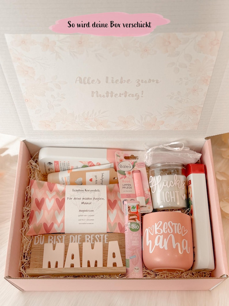 Może przedstawiać: R&oacute;żowe pudełko prezentowe wypełnione produktami do pielęgnacji. Zawiera kubek z napisem "BESTE MAMA", drewniany znak z napisem "DU BIST DIE BESTE MAMA" i inne produkty kosmetyczne. Pudełko jest ozdobione kwiatowymi wzorami i niemieckim tekstem.