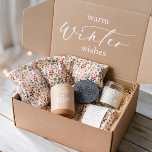 Könnte beinhalten: Eine Geschenkschachtel aus Karton mit dem Text warm winter wishes auf dem Innendeckel. Die Box enthält ein Wärmekissen mit Blumenmuster, eine beigefarbene Kerze mit dem Text time to relax, einen dunkelgrauen Untersetzer und eine Kerze.