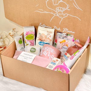 Könnte beinhalten: Eine braune Geschenkbox aus Pappe, gefüllt mit Baby-Pflegeprodukten. Die Box ist geöffnet und zeigt Artikel wie Lotionen, Cremes und Snacks. Der Deckel der Box hat eine handgezeichnete Herzform und die Worte "Entspann Dich, Mama".