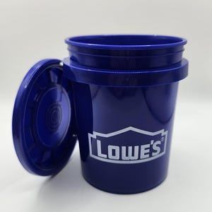Op de afbeelding: Een donkerblauwe plastic emmer met bijpassende deksel, met het Lowe's logo in wit. De deksel is iets geopend, waardoor de binnenkant zichtbaar is. Geschikt voor diverse huishoudelijke en bouwklussen.