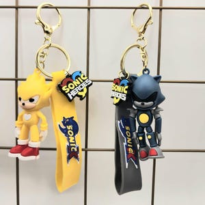 May include: Two keychains featuring Sonic the Hedgehog characters. One keychain has a yellow figure with red shoes and a yellow strap with the Sonic logo. The other keychain has a blue robot figure with a gray strap. Both have gold-colored key rings.