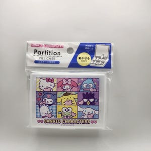 Puede incluir: Un pastillero blanco de Sanrio Characters en un embalaje transparente. El estuche presenta una cuadrícula de personajes de dibujos animados coloridos. El embalaje incluye texto en inglés y japonés. El pastillero está diseñado con separaciones móviles.
