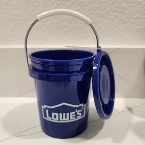 May include: A small, dark blue bucket with a white handle and a matching lid. The bucket has the Lowe's logo printed on the front. The lid is partially open, revealing the interior of the bucket.