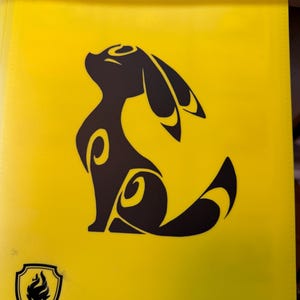 May include: A yellow binder featuring a black silhouette of a stylized creature with large ears and a curved tail. A shield emblem with a flame design is in the lower left corner. The binder is made of a smooth, glossy material.