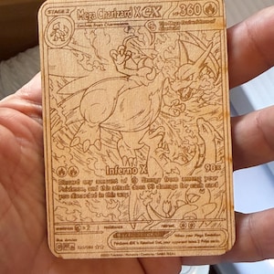 May include: A wooden Mega Charizard X&X Pokémon card. The card features an engraved illustration of Charizard in a dynamic pose, with the text "Inferno X" and other game details. The card is rectangular with rounded corners.