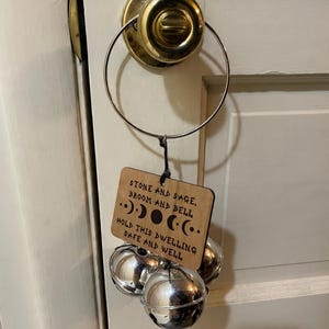Op de afbeelding: Een houten plaat met tekst en drie zilveren bellen die aan een deurklink hangen. De plaat leest "STONE AND SAGE, BROOM AND BELL, HOLD THIS DWELLING SAFE AND WELL." De bellen zijn bolvormig en glanzend.