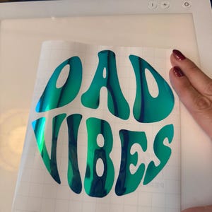 Pode incluir: Um autocolante de vinil com as palavras "BAD VIBES" num design em espiral azul e verde. O autocolante está sobre um fundo de grelha branca, com uma fonte arredondada.