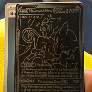 May include: Black and gold Mewtwo & Mew GX Tag Team Pokémon trading card. The card features a stylized illustration of the two Pokémon. The card has the text "BASIC Mewtwo & Mew GX HP 270 TAG TEAM".