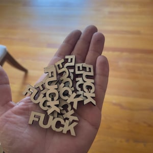 Puede incluir: Una mano sosteniendo un montón de pequeñas letras de madera recortadas que deletrean la palabra "FUCK". Las letras son de color madera natural y están esparcidas por la palma.