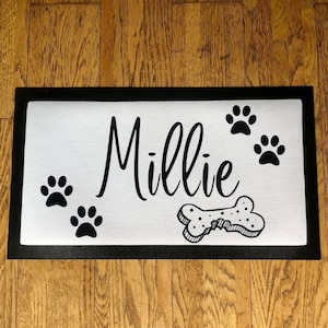 May include: Black and white doormat with the name "Millie" and paw prints. A bone-shaped treat is also on the mat.