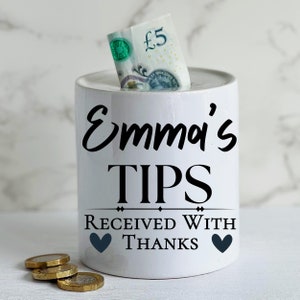 May include: White ceramic money box with black text that reads "Emma's TIPS RECEIVED WITH THANKS" and two black hearts. A five pound note is inserted into the slot on top of the box.