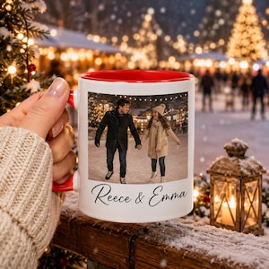May include: A white ceramic mug with a bright red interior and handle, featuring a printed photograph of two people ice skating outdoors and the black cursive text "Reece & Emma" printed below the image on the front of the mug.