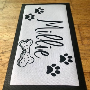 May include: Black and white doormat with the name "Millie" written in cursive. The doormat also features a bone and paw prints.