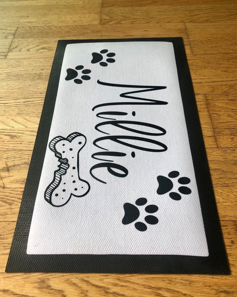 May include: Black and white doormat with the name "Millie" written in cursive. The doormat also features a bone and paw prints.