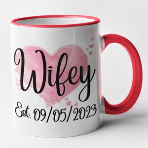 May include: White ceramic mug with a red rim and handle. The mug features a pink watercolor heart with the word "Wifey" in black script. The date "Est. 09/05/2023" is written below the heart.